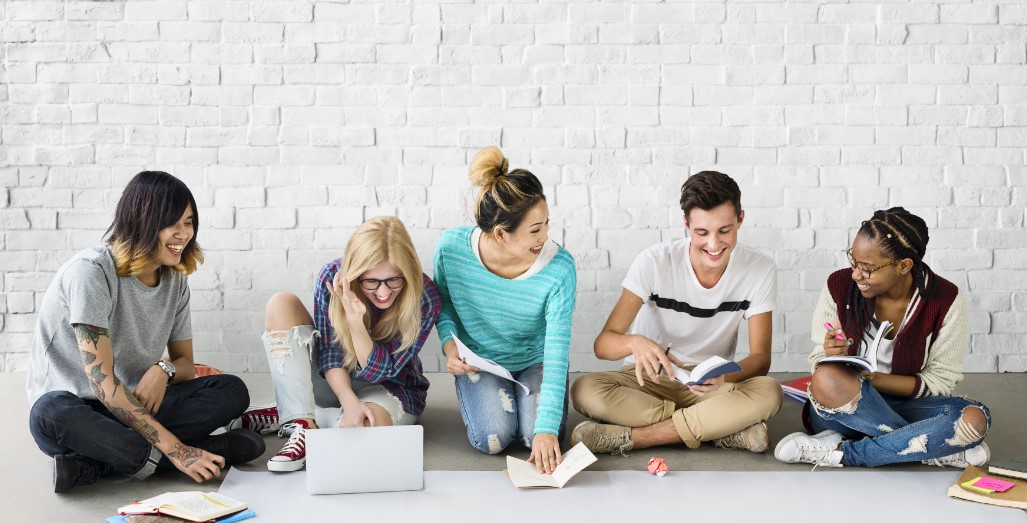 Fünf junge Personen sitzen im Halbkreis auf dem Boden mit Büchern und Laptop und lernen gemeinsam.